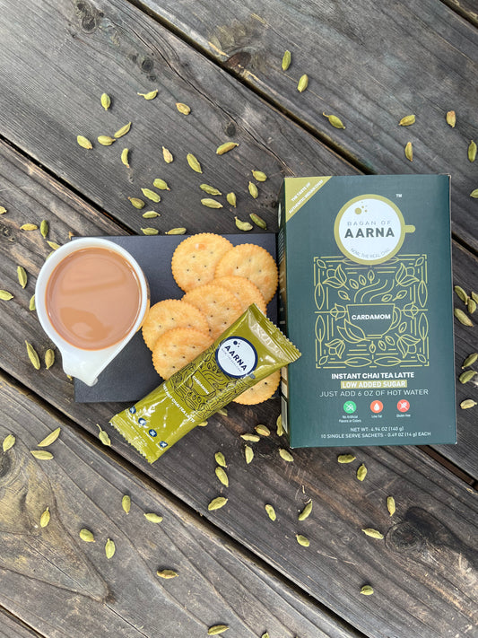 A box of Aarna Instant Cardamom Chai Tea next to a cup of tea with milk and biscuits, all placed on a wooden surface with cardamom seeds scattered around.